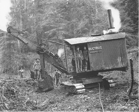 A black and white photo of men posing with a steam shovel. Before vehicles could become autonomous, they had to become mechanized. Two men are standing in the sheet metal cab of the shovel and one is standing on the bucket. The shovel is parked among downed trees with a forest in the background