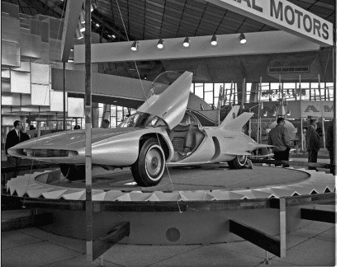 General Motors' autonomous vehicle concept, the Firebird III, sits on a display plinth at a prototype car show. The driver's side door is open and its unique hinges cause the door to open upward rather than out. The vehicle has airplane-like fins on the rear and sensors on the bottom to follow a guide wire embedded in the road